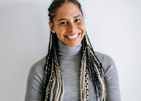 Smiling ethnic female with long braids in turtleneck and jeans crossing hands standing against white background